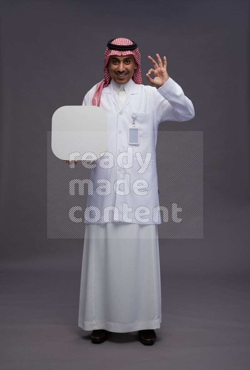 Saudi man wearing thob with lab coat and shomag with pocket employee badge standing holding social media sign on gray background