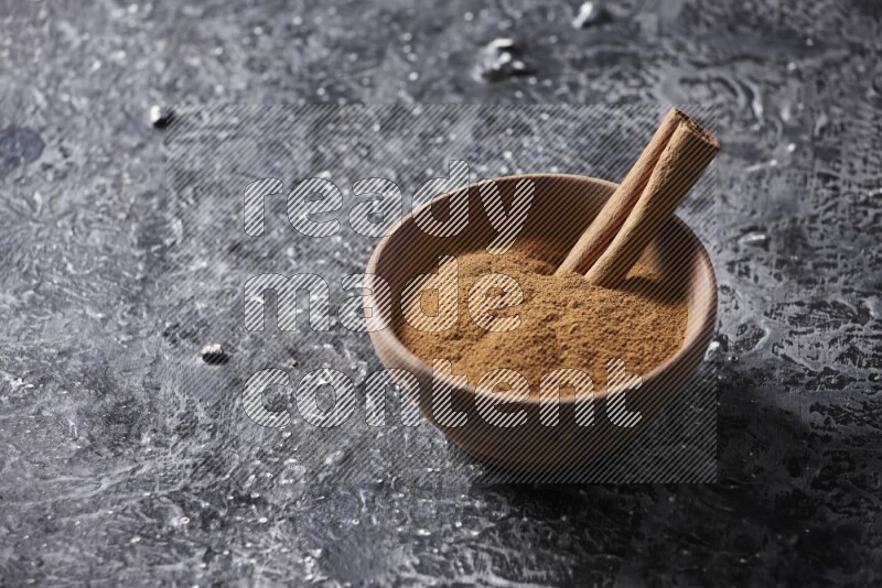 Wooden bowl full of cinnamon powder and a cinnamon stick on a textured black background