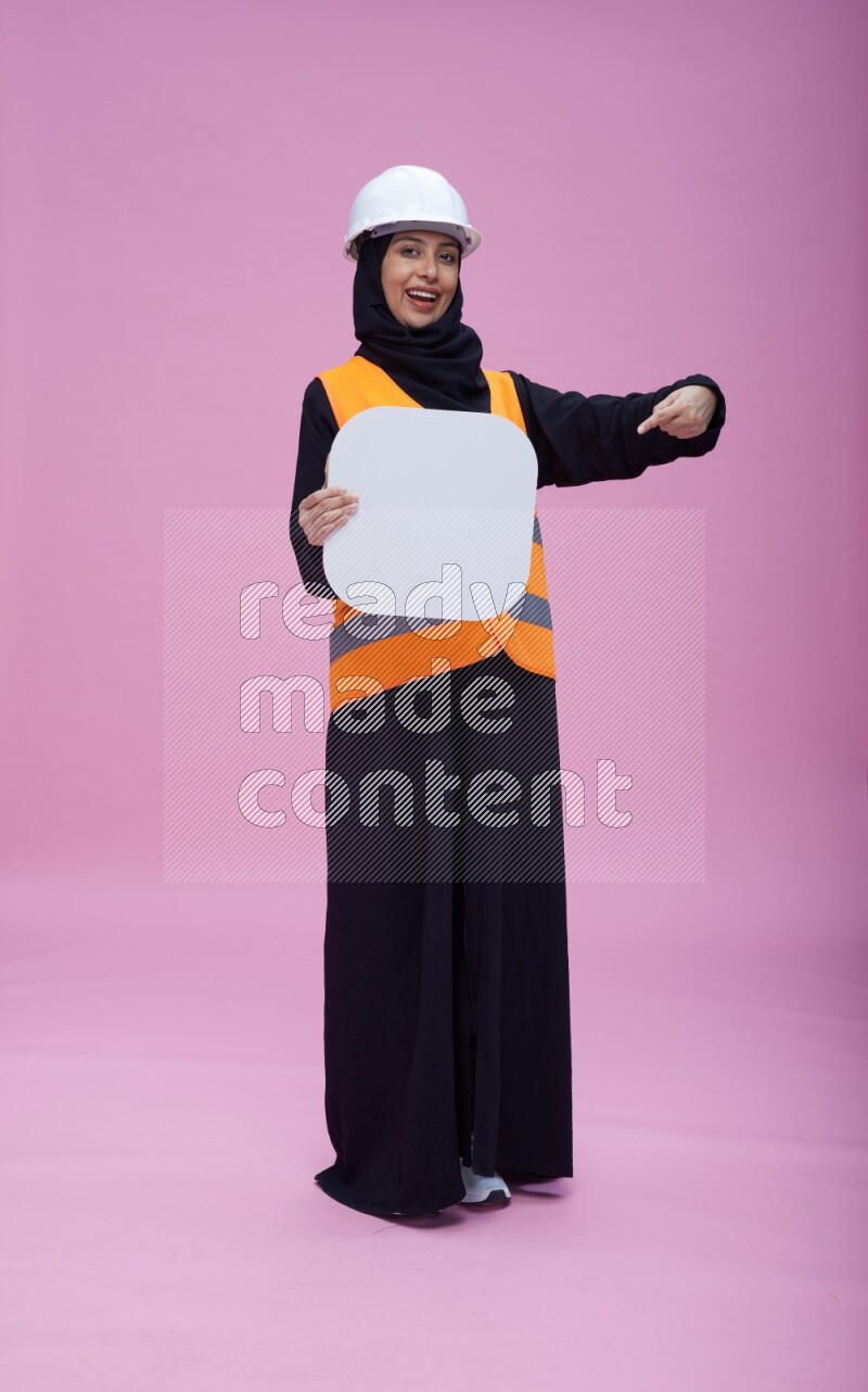 Saudi woman wearing Abaya with engineer vest and helmet standing holding social media sign on pink background