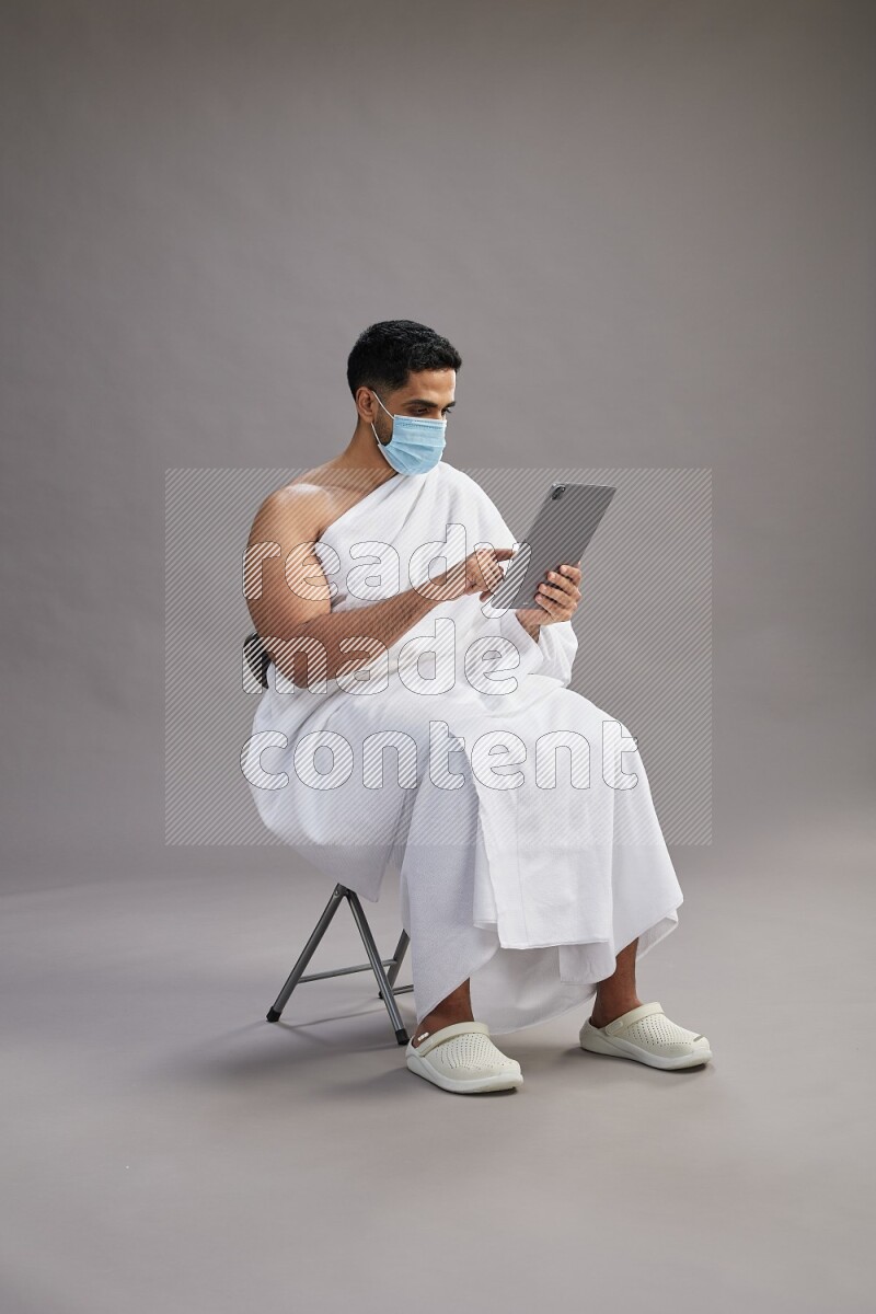 A man wearing Ehram with face mask sitting on chair holding a tablet on gray background