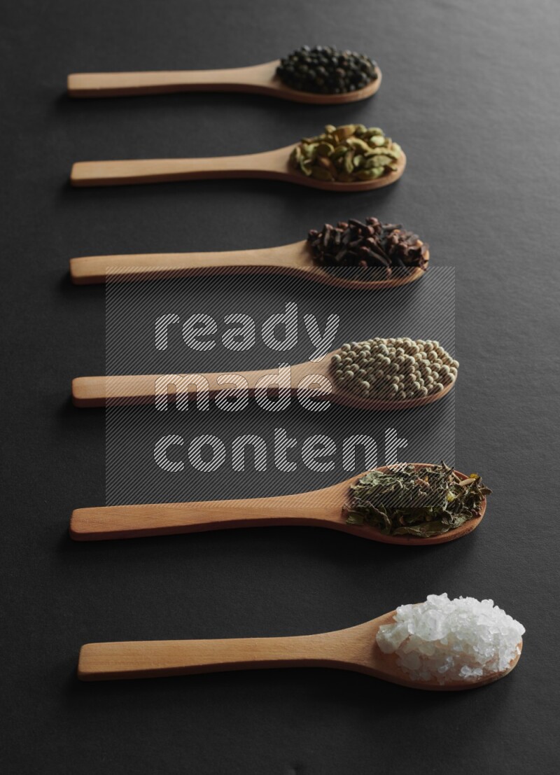 wooden spoons filled with white peppers, cloves, cardamom, salt, black peppers and basil on black flooring