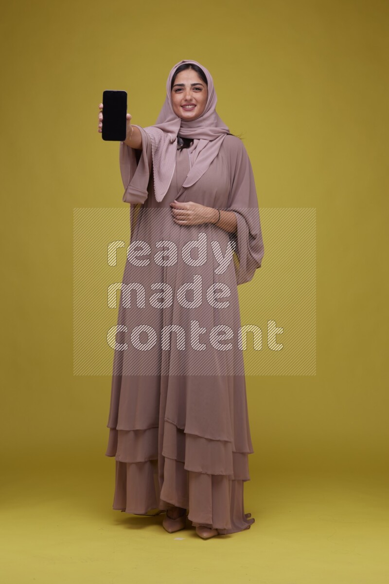 A woman Showing her Phone screen on a Yellow Background wearing Brown Abaya with Hijab