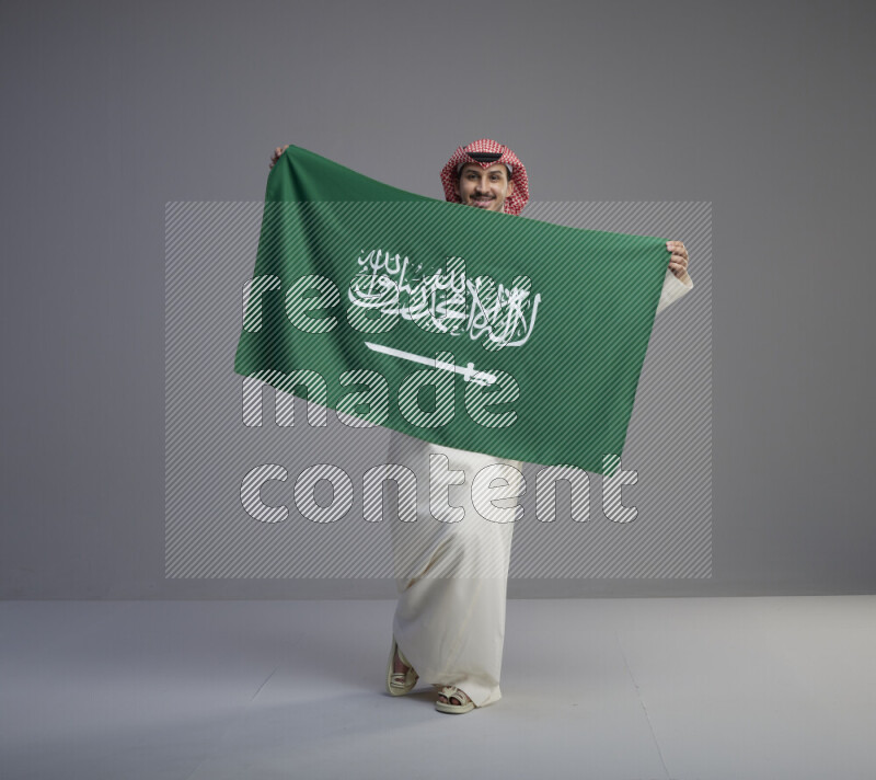 A saudi man standing wearing thob and red shomag holding big saudi flag on gray background