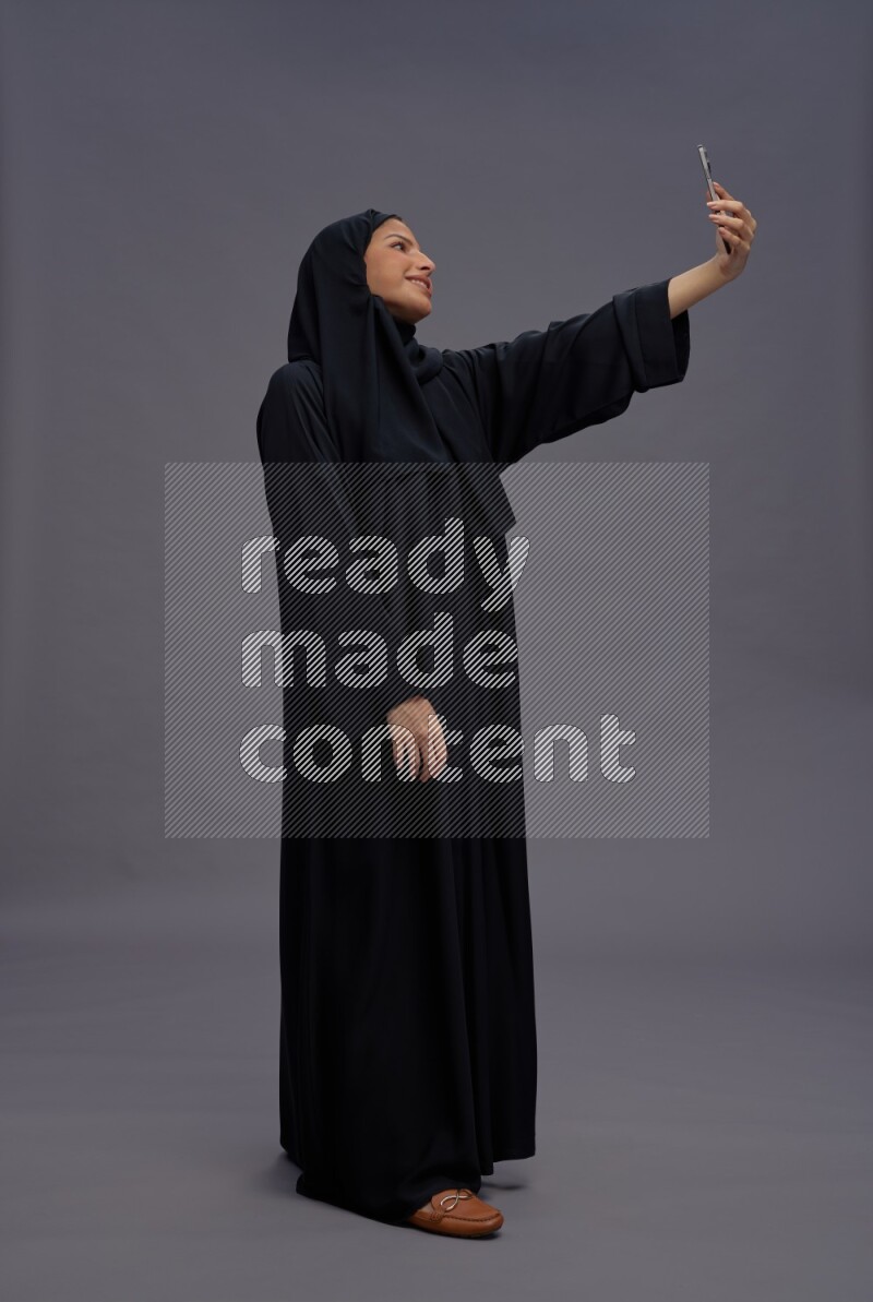 Saudi woman wearing Abaya standing taking selfie on gray background