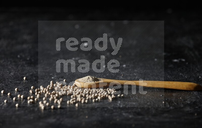 A wooden spoon full of white pepper powder with white pepper beads on textured black flooring