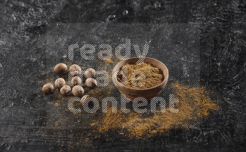 A wooden bowl full of nutmeg powder with the seeds and sprinkled powder beside it on a textured black flooring in different angles