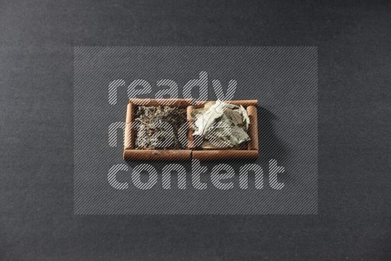 مربعان من أعواد القرفة ممتلئان بأوراق الغار والريحان المجفف علي خلفية سوداء