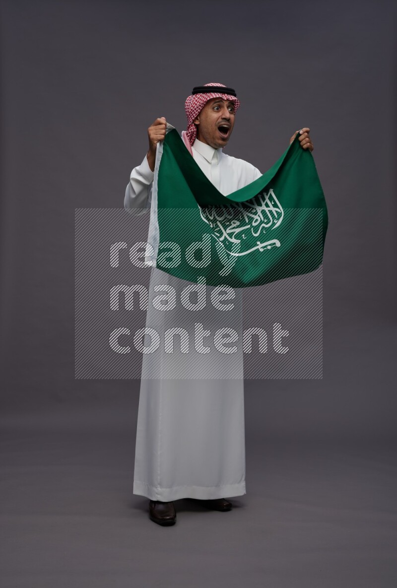 Saudi man wearing thob and shomag standing holding Saudi flag on gray background