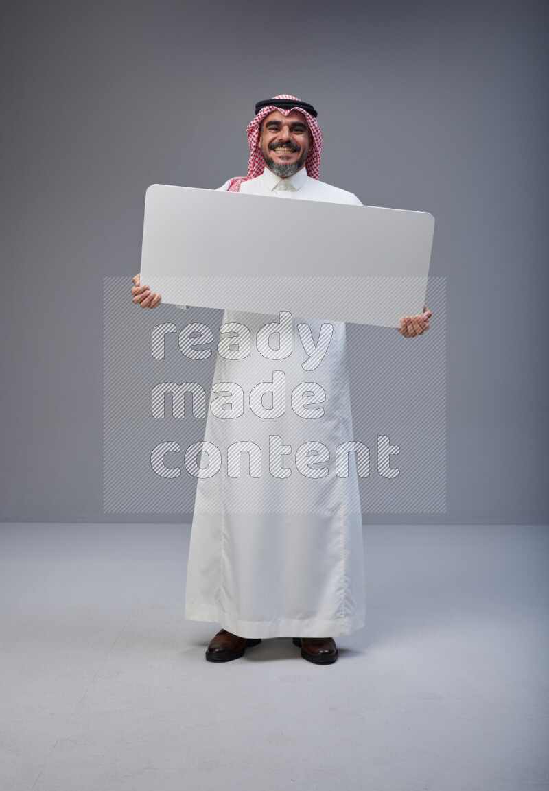 Saudi man Wearing Thob and red Shomag standing holding board on Gray background