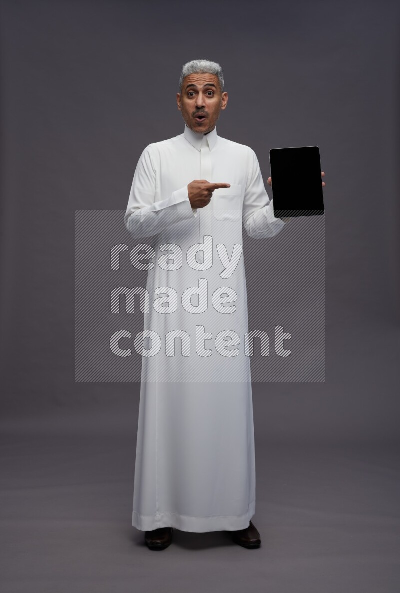 Saudi man wearing thob standing showing tablet to camera on gray background