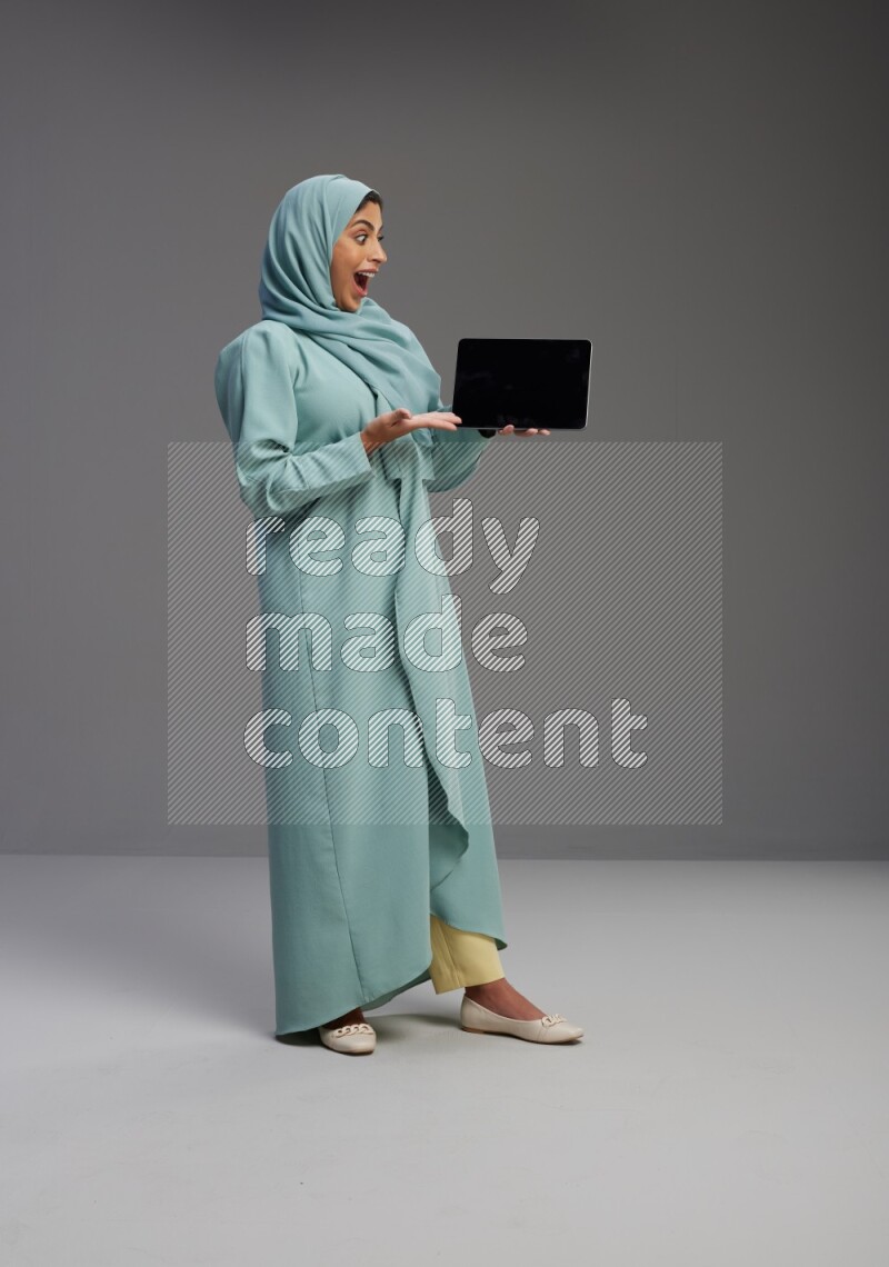 Saudi Woman wearing Abaya standing showing tablet to camera on Gray background