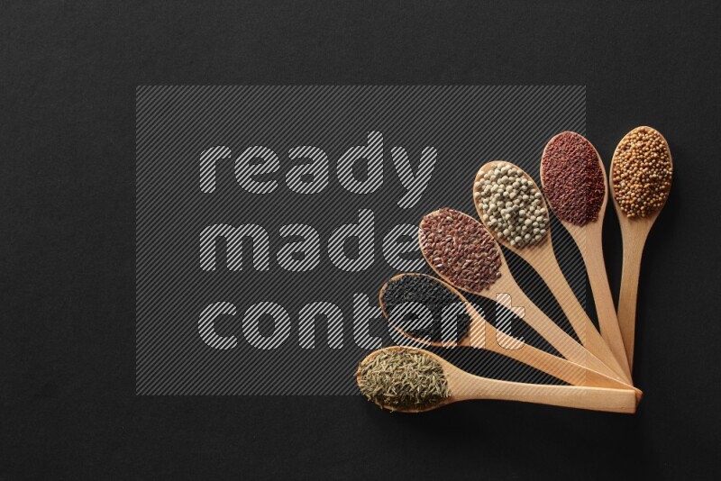 wooden spoons filled with white peppers, mustard seeds, black seeds, garden cress, cumin and flax on black flooring