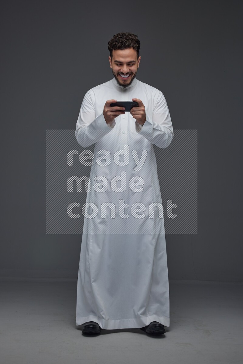 A Saudi man wearing Thobe and playing using his phone horizontally playing video games while standing and making different poses eye level on a gray background