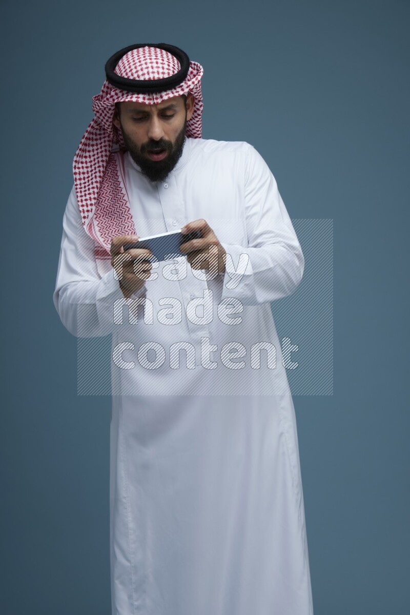 A man Playing a Game on his phone in a blue background wearing Saudi Thob with Shomag