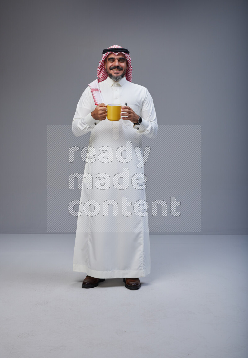 Saudi man Wearing Thob and red Shomag standing holding mug on Gray background