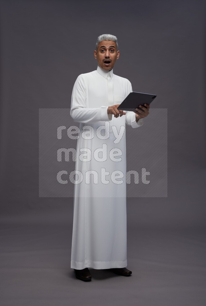 Saudi man wearing thob standing working on tablet on gray background