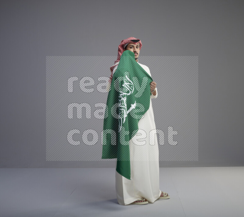 A saudi man standing wearing thob and red shomag holding big saudi flag on gray background