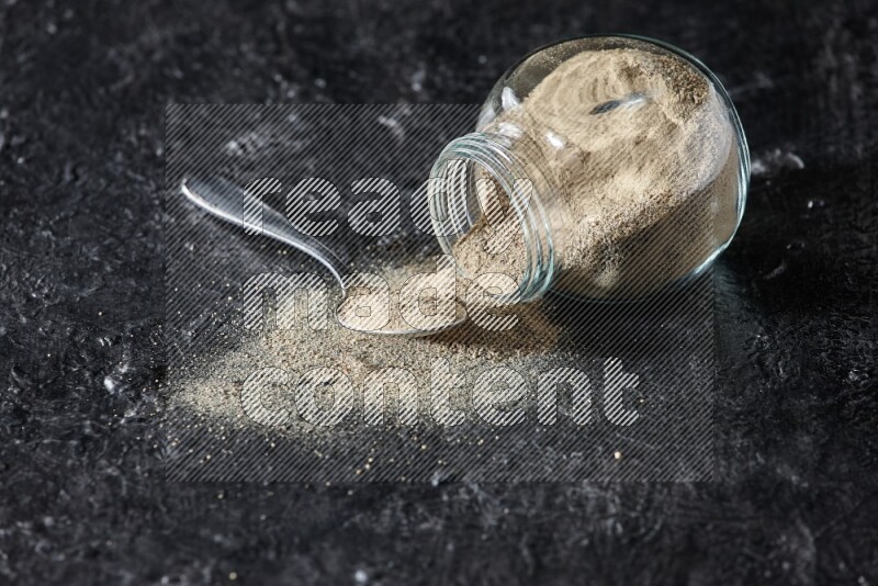 A flipped herbal glass jar and a metal spoon full of white pepper powder with spilled powder on textured black flooring