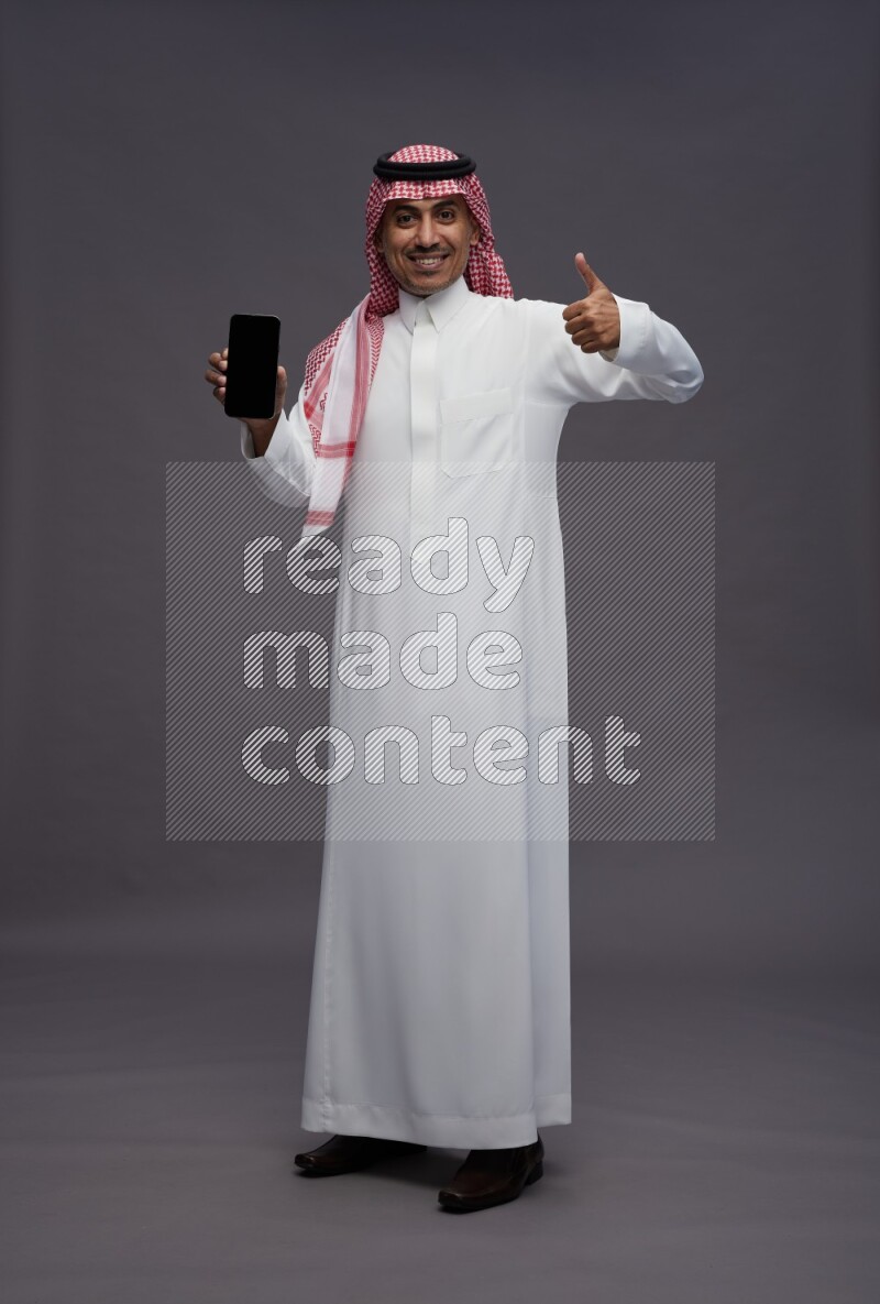 Saudi man wearing thob and shomag standing showing phone to camera on gray background