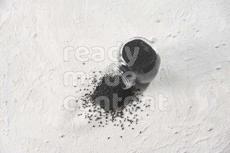 A glass spice jar full of black seeds and the jar flipped and seeds spread on a textured white flooring