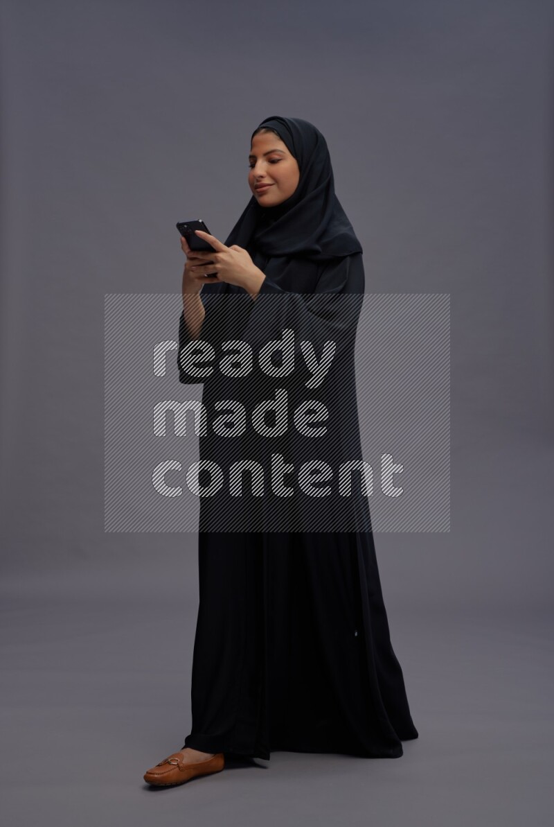 Saudi woman wearing Abaya standing texting on phone on gray background