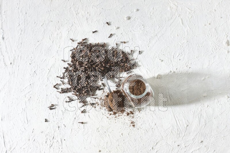 A glass spice jar and a metal spoon full of cloves powder and cloves spread on textured white flooring