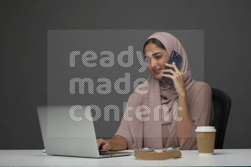 A Saudi woman Setting on her desk
 calling  on a Gray Background wearing Brown Abaya with Hijab
