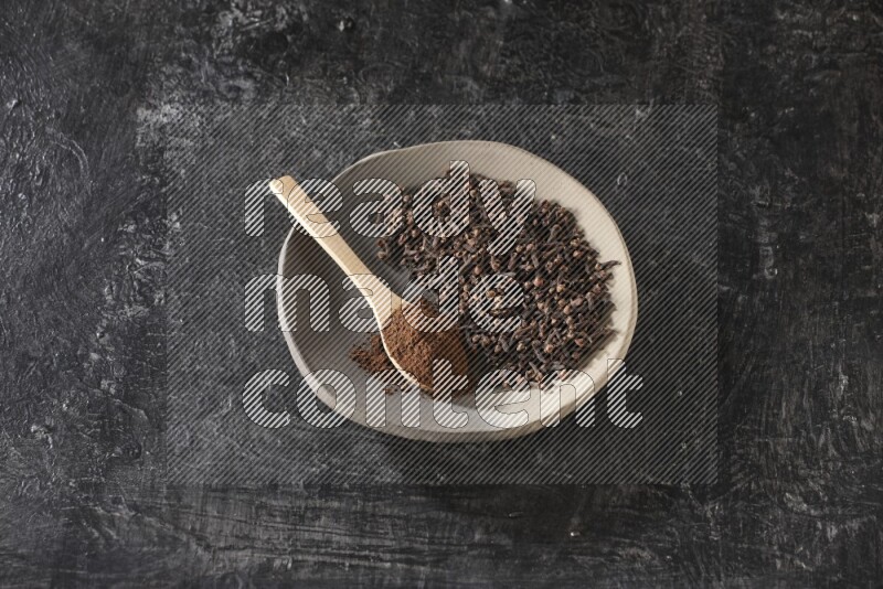 A Pottery plate full of cloves and a wooden spoon full of cloves powder on it on a textured black background