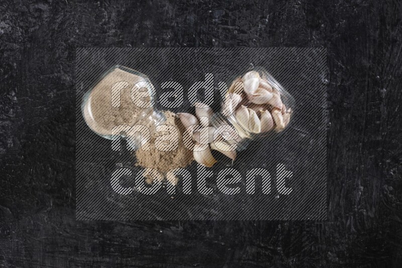 2 glass spice jars, one full of garlic powder and the other full of garlic cloves both flipped on a textured black flooring