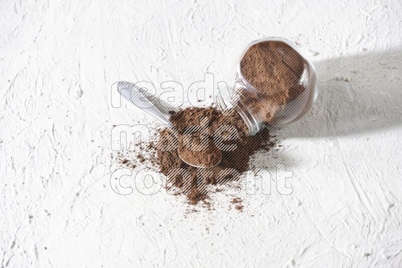 A flipped glass spice jar and a metal spoon full of cloves powder and powder came out of the jar on textured white flooring