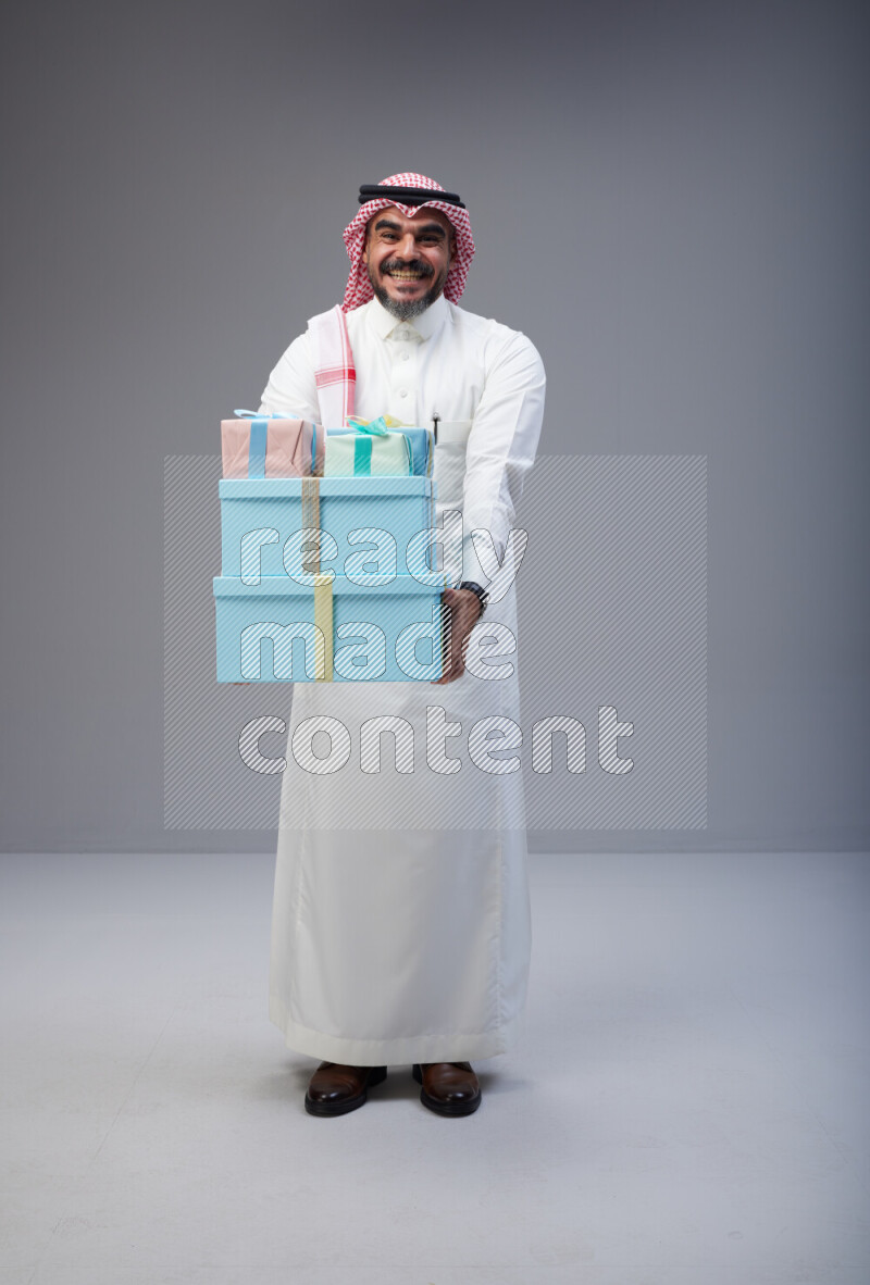 Saudi man Wearing Thob and red Shomag standing holding gift box on Gray background
