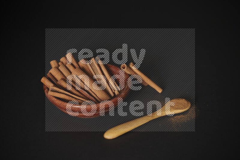 Cinnamon sticks in wooden bowl and cinnamon powder in a wooden spoon on black background
