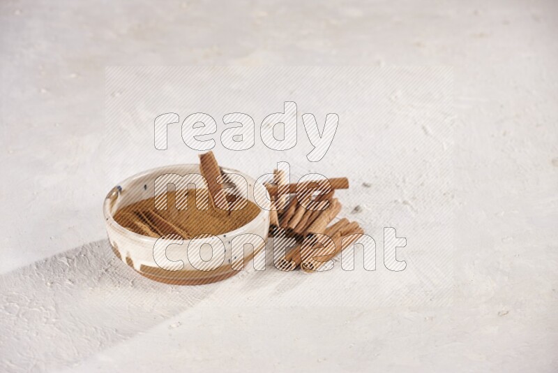 Ceramic bowl full of cinnamon powder with cinnamon sticks on the side on white background