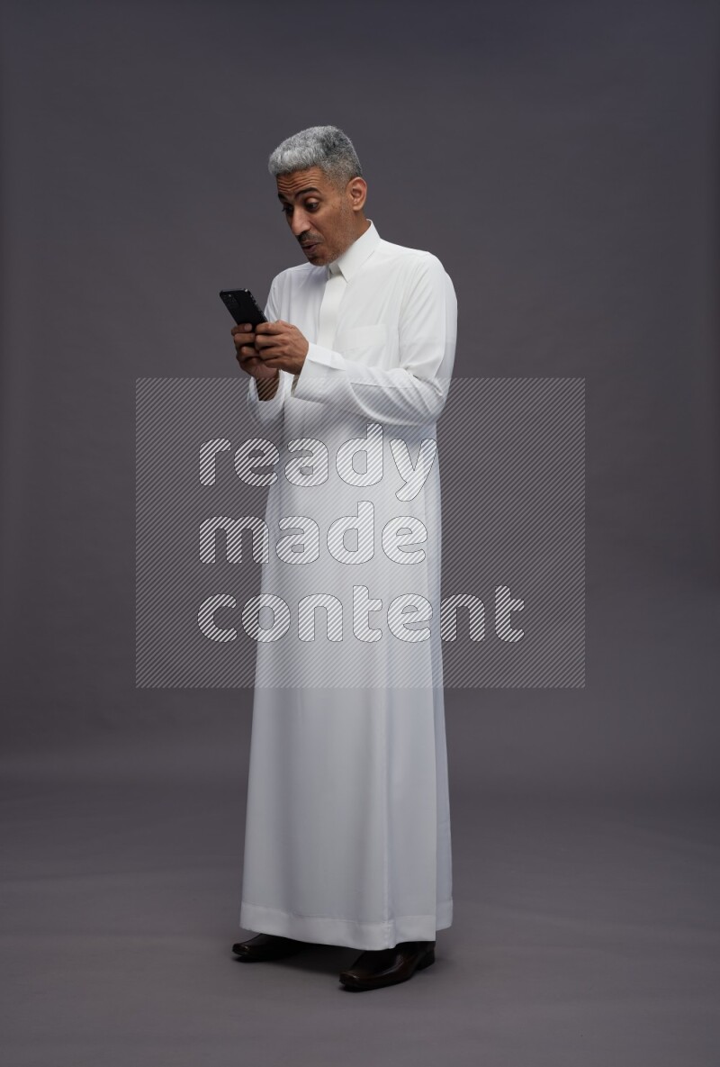 Saudi man wearing thob standing texting on phone on gray background