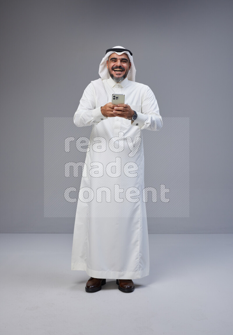 Saudi man Wearing Thob and white Shomag standing texting on phone on Gray background