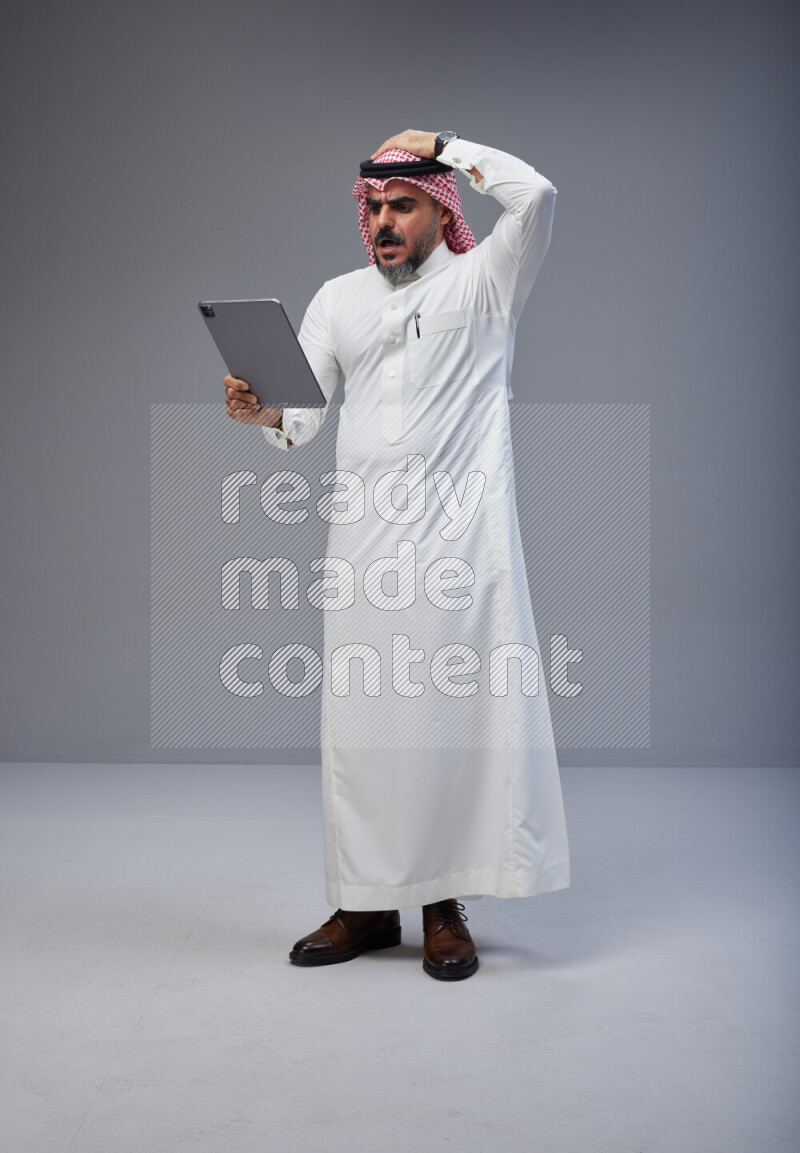 Saudi man Wearing Thob and red Shomag standing working on tablet on Gray background