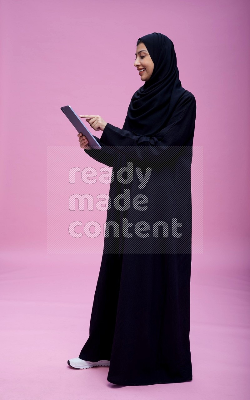 Saudi woman wearing Abaya standing working on tablet on pink background