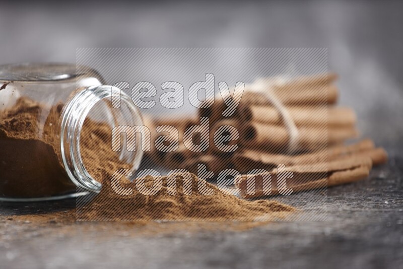 Herbal glass jar full of cinnamon powder flipped with cinnamon sticks stacked and bounded on a textured black background
