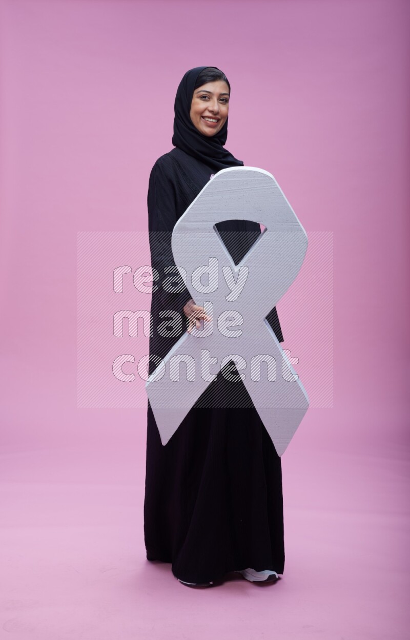 Saudi woman wearing pink ribbon on abaya standing holding awareness ribbon on plain pink background