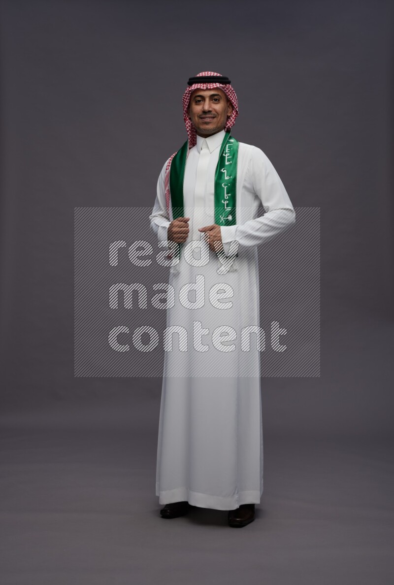Saudi man wearing thob and shomag standing holding Saudi flag on gray background