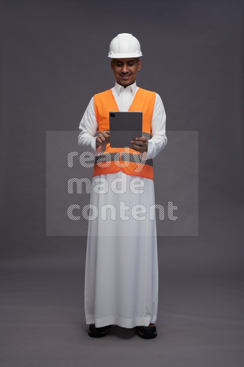 Saudi man wearing thob with engineer vest standing working on tablet on gray background