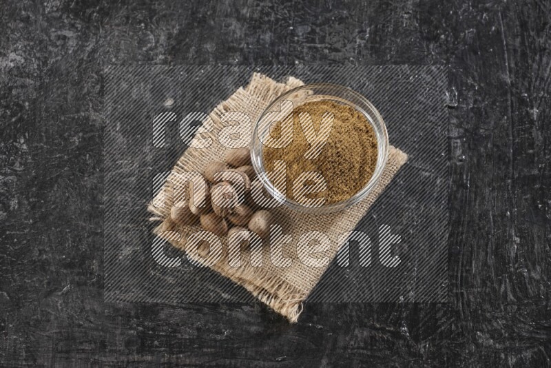 A glass bowl full of nutmeg powder with whole seeds beside it on burlap fabric on a textured black flooring