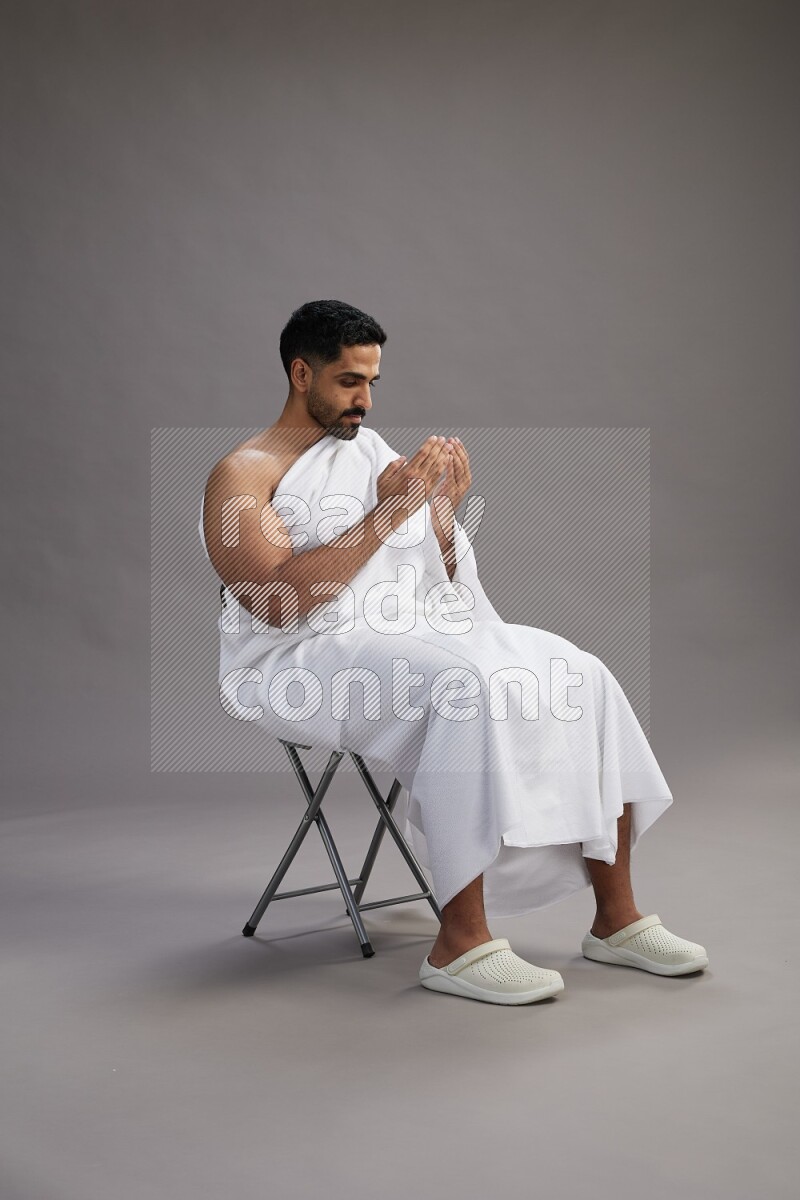 A man wearing Ehram sitting on chair performing dua'a on gray background