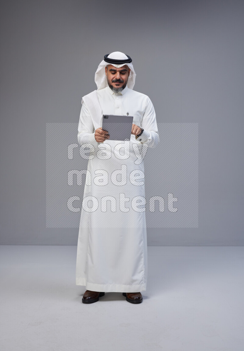 Saudi man Wearing Thob and white Shomag standing working on tablet on Gray background