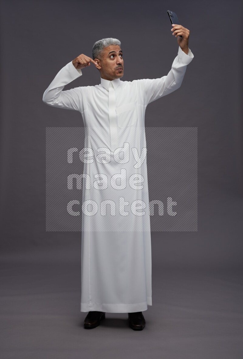 Saudi man wearing thob standing taking selfie on gray background