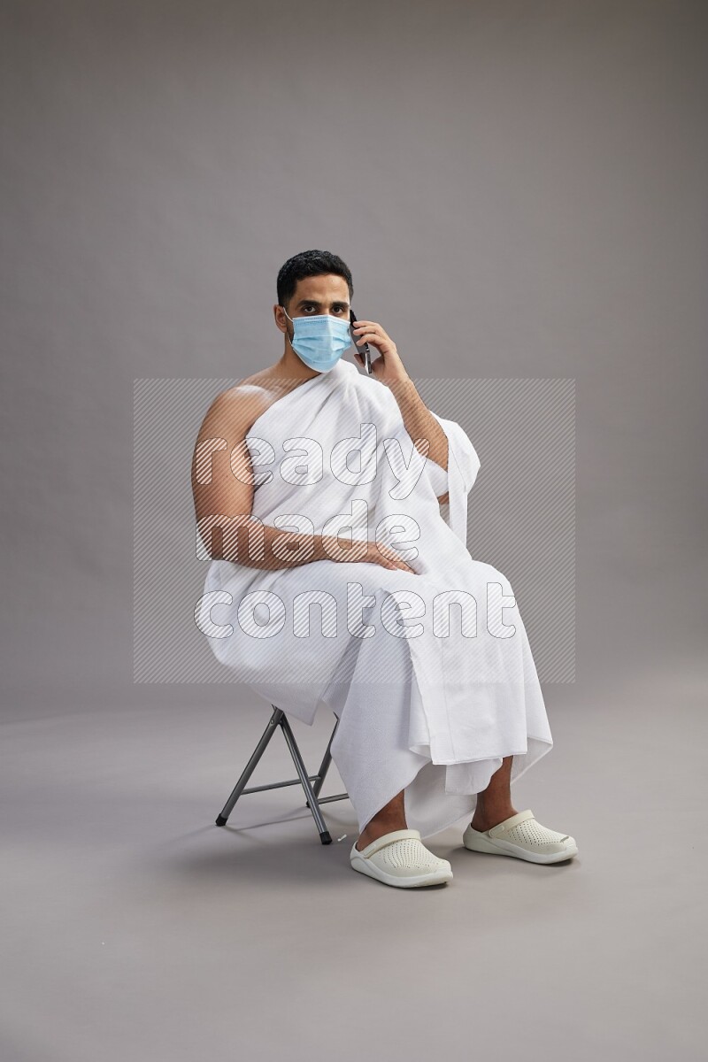 A man wearing Ehram with face mask sitting on chair talking on phone on gray background