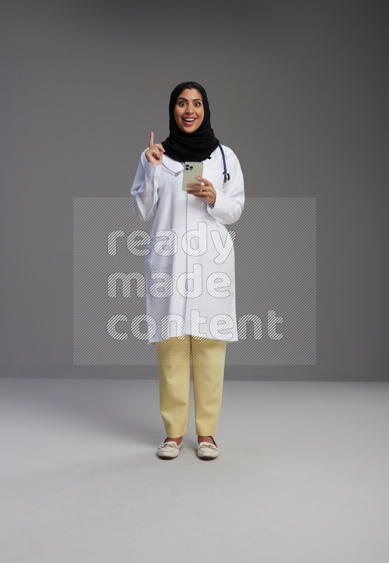 Saudi woman wearing lab coat with stethoscope standing texting on phone on Gray background