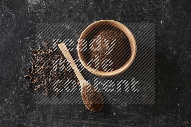 A wooden bowl and a wooden spoon full of cloves powder with spreaded cloves on a textured black flooring
