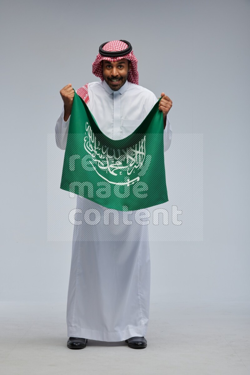 Saudi man Wearing Thob and shomag standing holding Saudi flag on Gray background