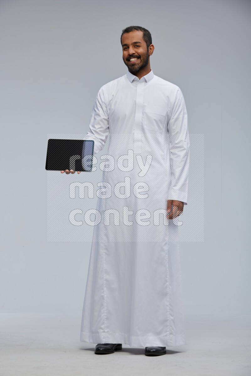 Saudi man wearing Thob standing showing tablet to camera on Gray background